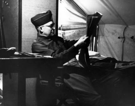Wearing glasses, Dwight Eisenhower reads a newspaper while sitting in a tent.