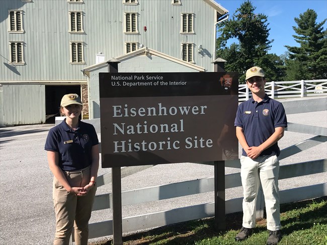 Two interns in blue shirts and khakis stand in front of a brown sign reading Eisenhower National Historic Site
