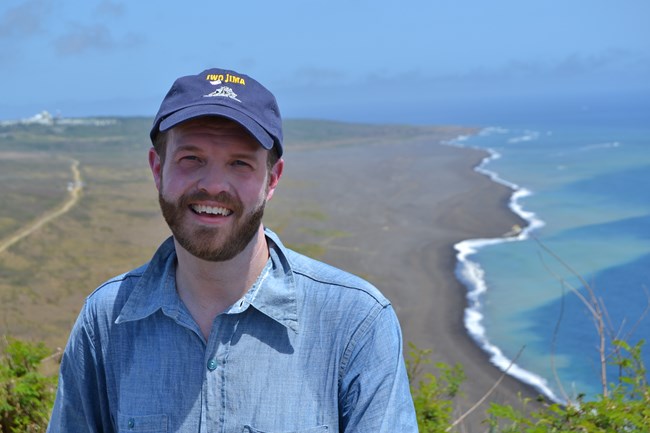 A man in a blue shirt stands atop a mountain with an ocean background