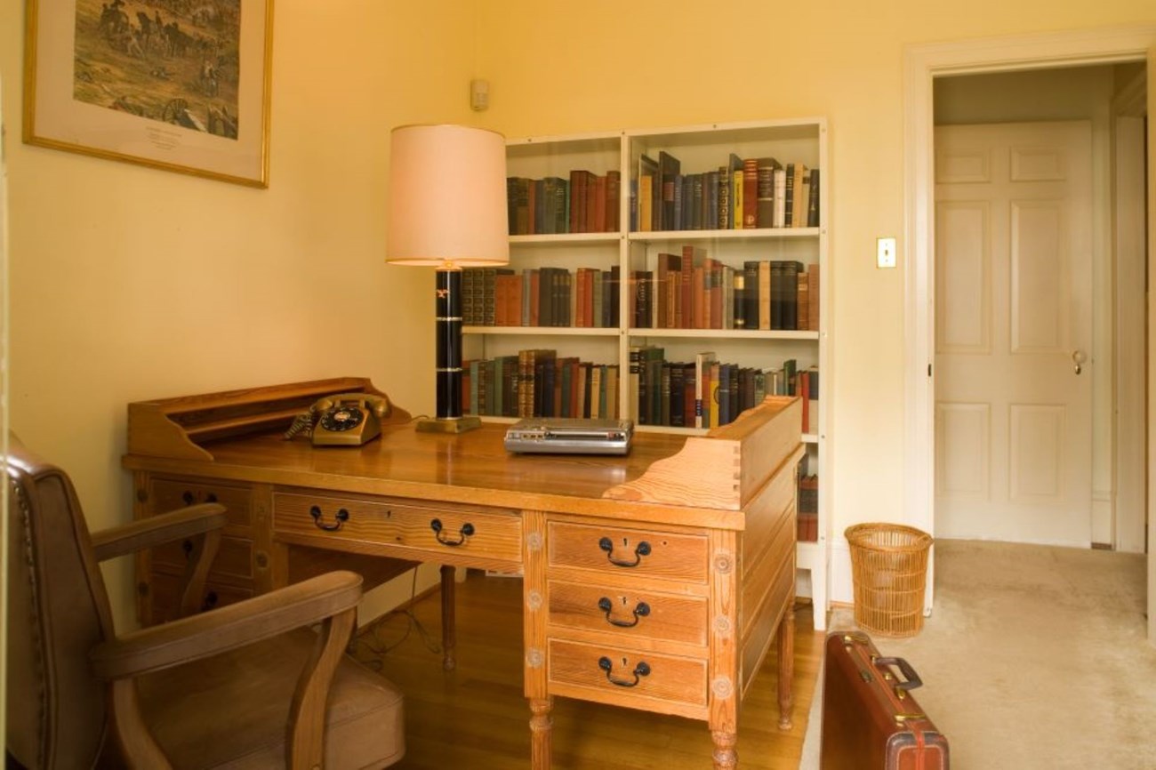 A wood desk sits in the middle of a small, yellow room, with a bookshelf on the other side