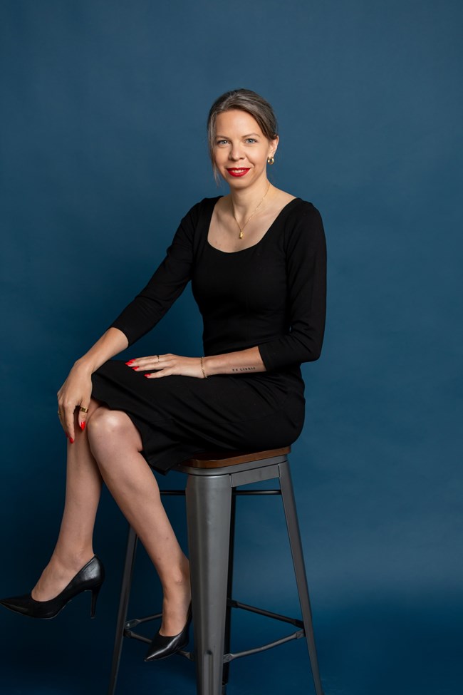 A woman in a dark dress sits on a stool witha  blue background