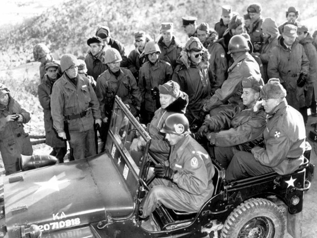 A black and white image showing President-elect Eisenhower wearing winter clothing and seated in a jeep, surrounded by army officers