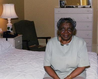 Delores Moaney, wearing a white dress, sits on the bed in the Moaney room at the Eisenhower home in Gettysburg. A lamp is on in the background. A rocking chair and dresser also stands in the background.