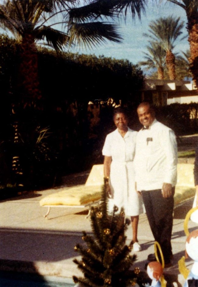 John and Delores Moaney in California ca. 1967 Delores and John Moaney, both wearing white, stand and smile for a photograph. In the background are some palm trees and a large building.