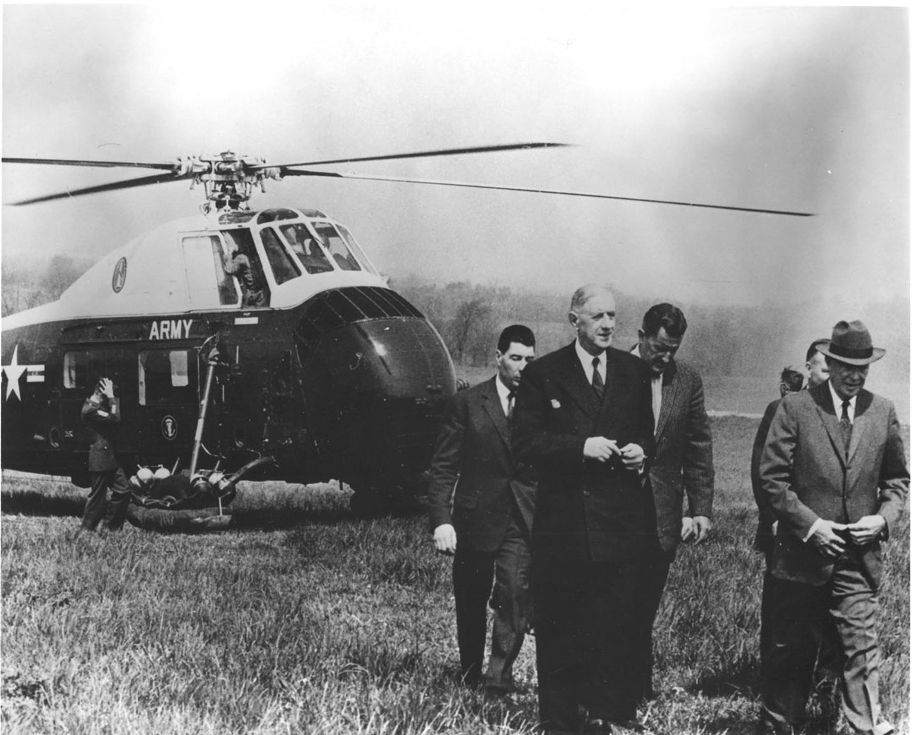 Dwight Eisenhower, wearing a grey suit and hat, walks alongside French President Charles DeGaulle, who has just arrived by helicopter to the Eisenhower Farm. Several other men in suits are in photograph.