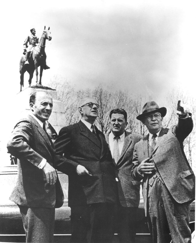 Dwight Eisenhower points toward the distance while Charles DeGaulle looks on; the two men, along with two others, stand in front of the large Virginia Memorial on the Gettysburg Battlefield