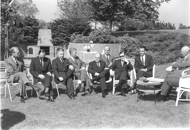 Congressman Gerald Ford sits with legs crossed on far left of photograph, while Dwight Eisenhower sits far right. Between them are a number of congressmen, while in the background is the brick patio and grill near the Eisenhowers' tea house