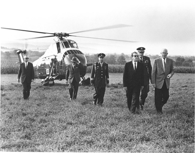 Dwight Eisenhower, wearing a grey suit, welcomes President Mateos to his farm. In the background is the large Marine One helicopter and several men wearing suits.