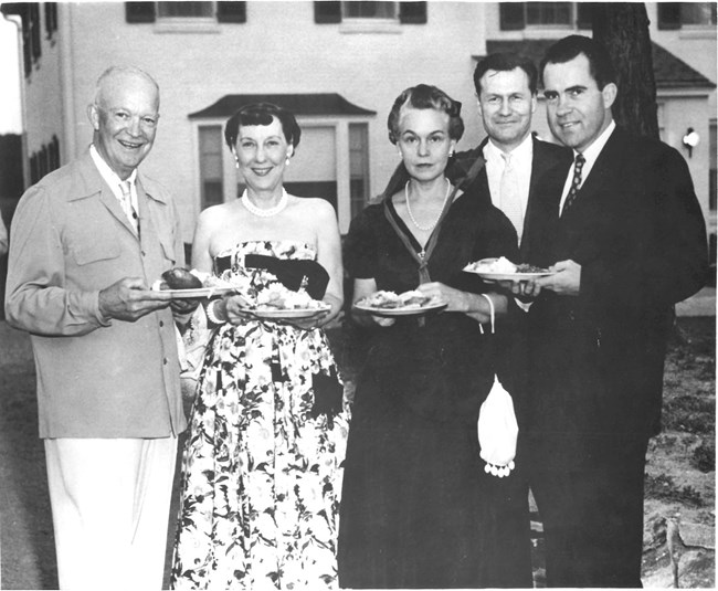 Dwight and Mamie stand with Ovetta Culp Hobby, Nelson Rockefeller, and Richard Nixon, in front of the white brick Eisenhower home, all holding plates of food