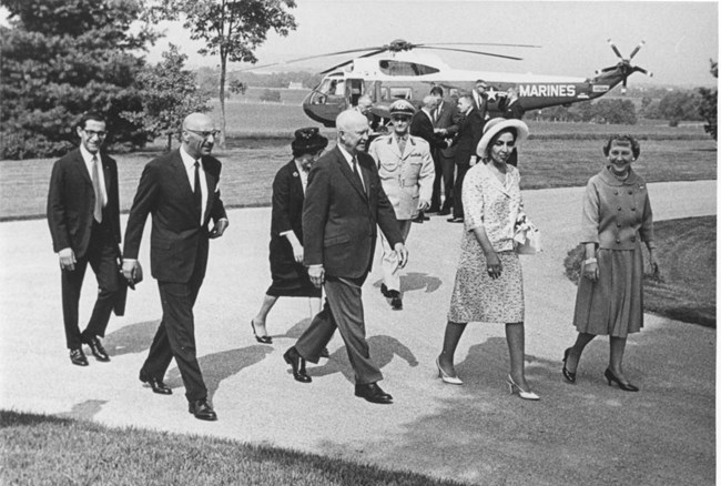 With the large Marine One Helicopter in the background, Dwight and Mamie Eisenhower walk with King Mohammed Shah Zahir and Queen Homeira Begum on the asphalt walkway toward the front door of the Eisenhowers' home. Several other men in suits are walking