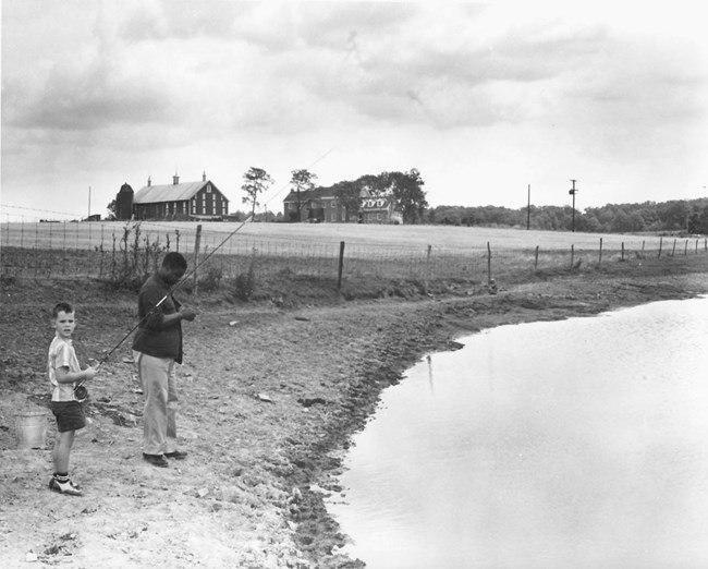 John Moaney Fishing With David Eisenhower A young David Eisenhower stands with John Moaney on the banks of a pond on the Eisenhower property; both are holding fishing rods. A home stands in the background.