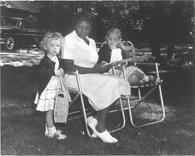 A seated Delores Moaney, wearing a white dress, reads a book to the young granddaughters of Dwight and Mamie Eisenhower