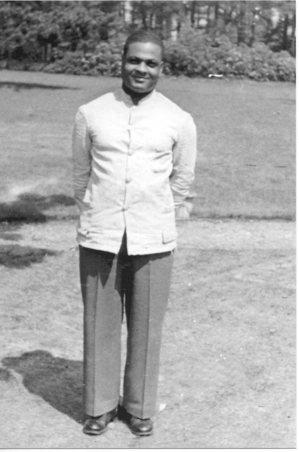 A smiling John Moaney stands for a photograph; he is wearing a white shirt and grey pants