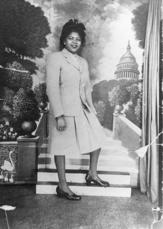 Delores Moaney stands for a photograph; she is wearing a dress. In the background is an image of the US Capitol Building