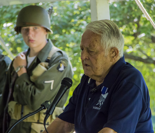A World War II author/historian speaks to a crowd while a living historian dressed in a United States World War II soldier looks on.