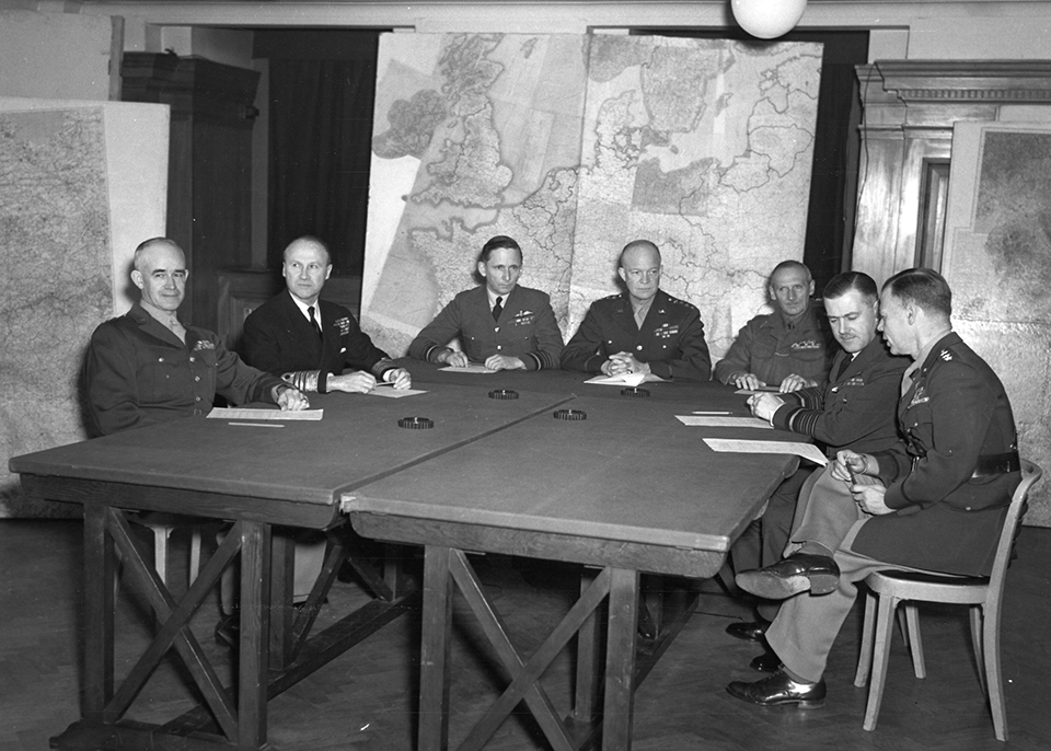 General Dwight D. Eisenhower and his commanders plan D-Day. A black and white photo of seven American military officers in full uniform sit around a table discussing military operations. There are multiple large maps in the background.