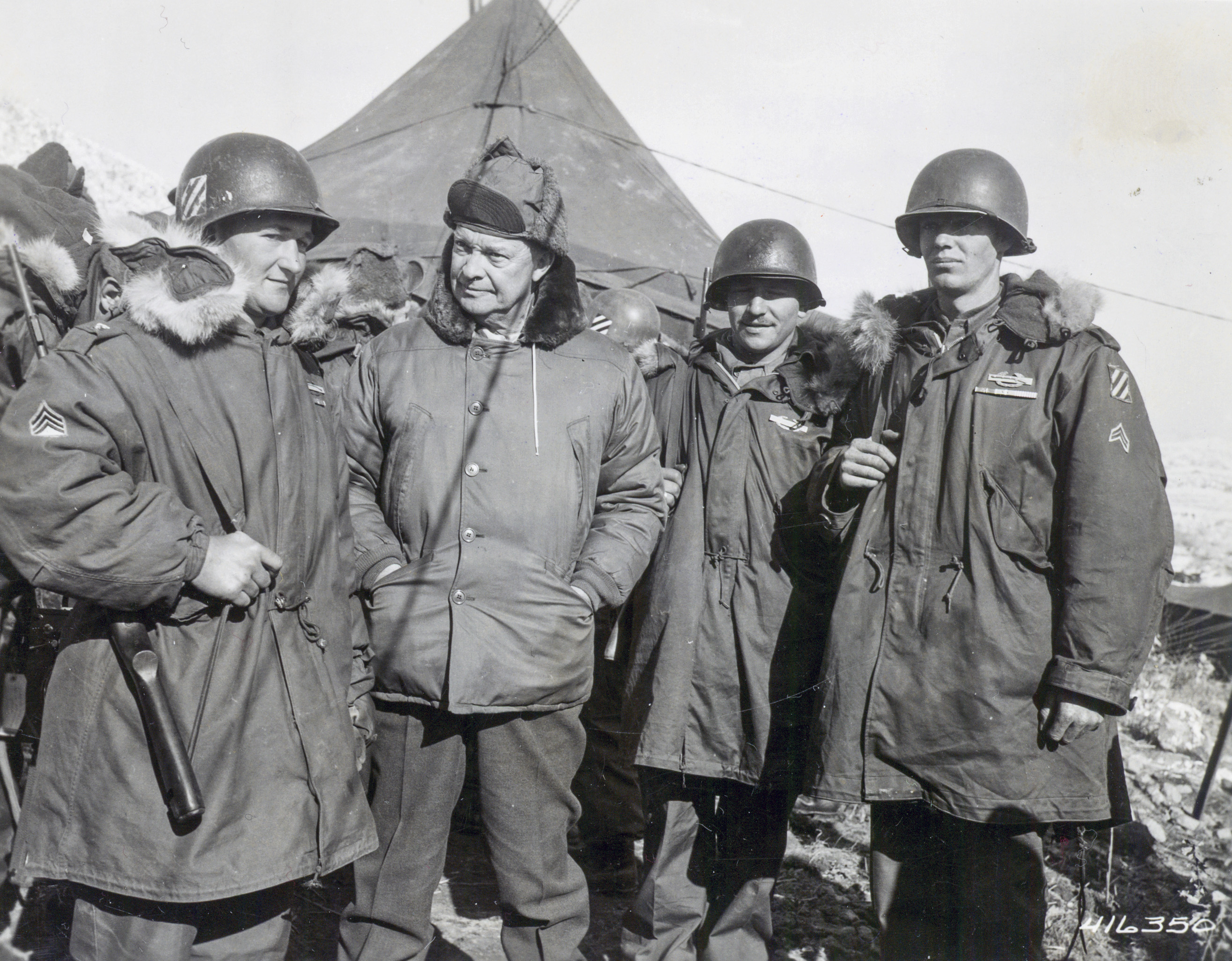 A black and white picture of Dwight Eisenhower standing with three soldiers during the Korean War. All four men are wearing heavy winter coats and are standing in from of a tent.