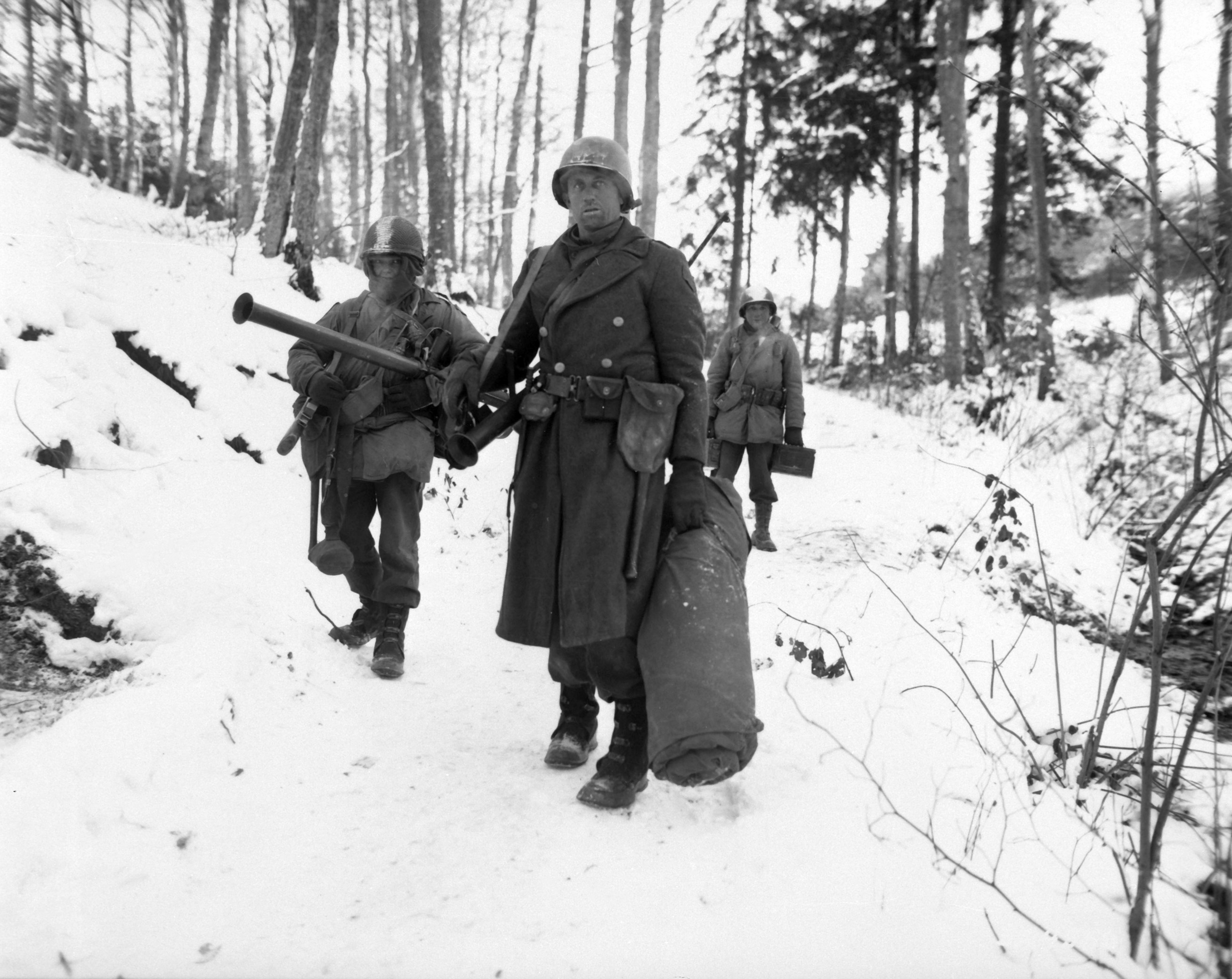 American engineers emerge from the woods and move out of defensive positions after fighting in the vicinity Bastogne, Belgium. American World War II soldiers emerge from the woods. They are dressed in heavy coats and carry large sacks. There is snow on the ground.