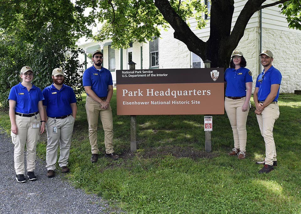 Five interns stand next to the park headquarters sign. They are standing under a large tree with a white building behind them. They are all wearing blue shirts and khaki pants and ball caps.