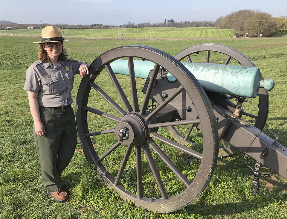 A female National Park Service ranger stands next to a Civil War cannon in an open green field.