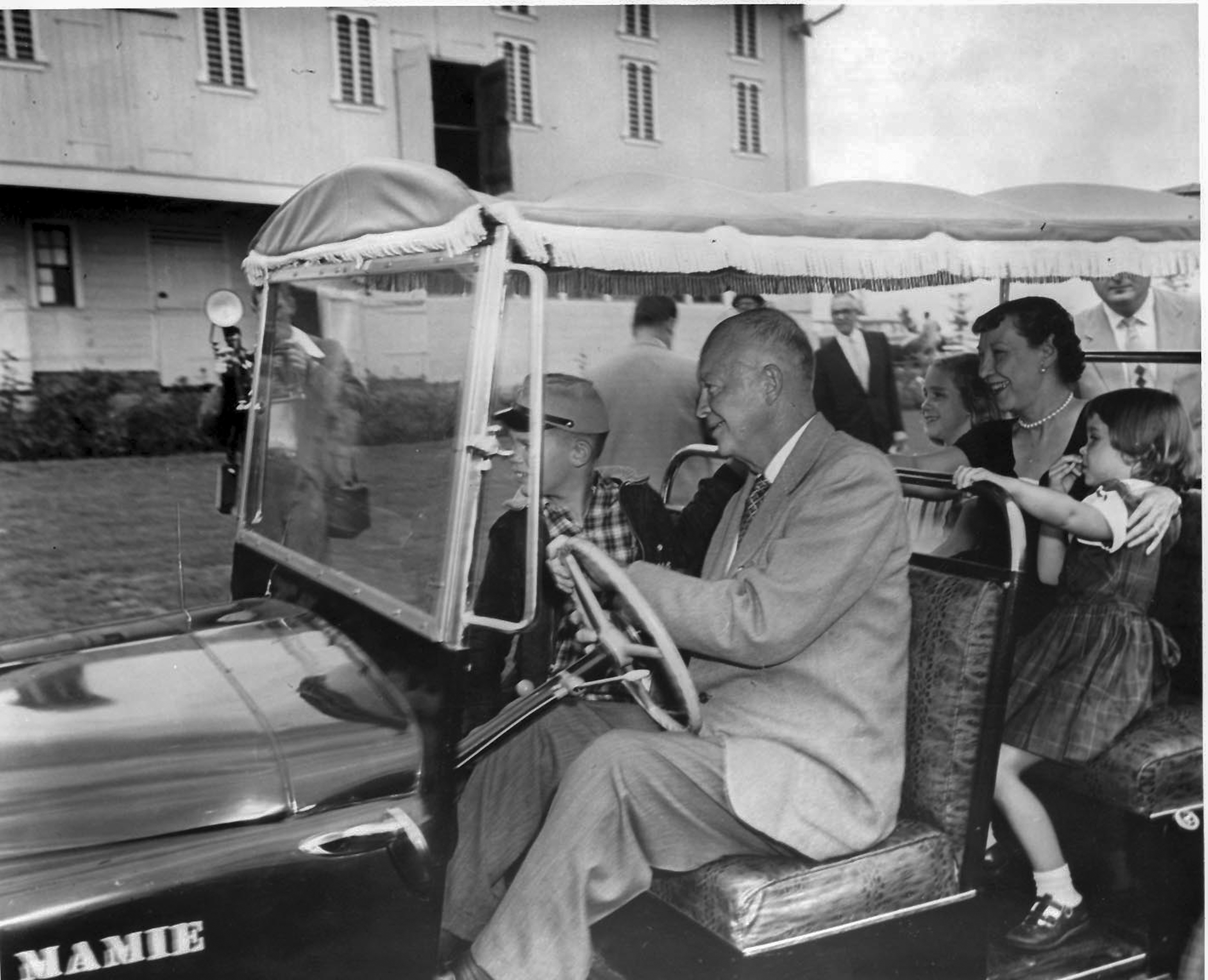 A black and white photo of President Dwight D. Eisenhower driving a golf cart with his wife Mamie and their grandchildren around their Gettysburg farm.