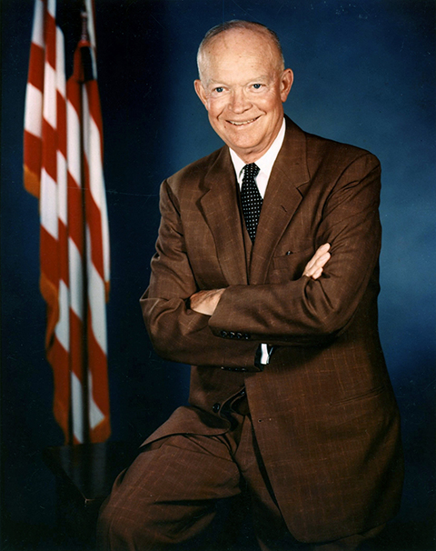 President Dwight D. Eisenhower, wearing a brown suit, stands next to the American flag.