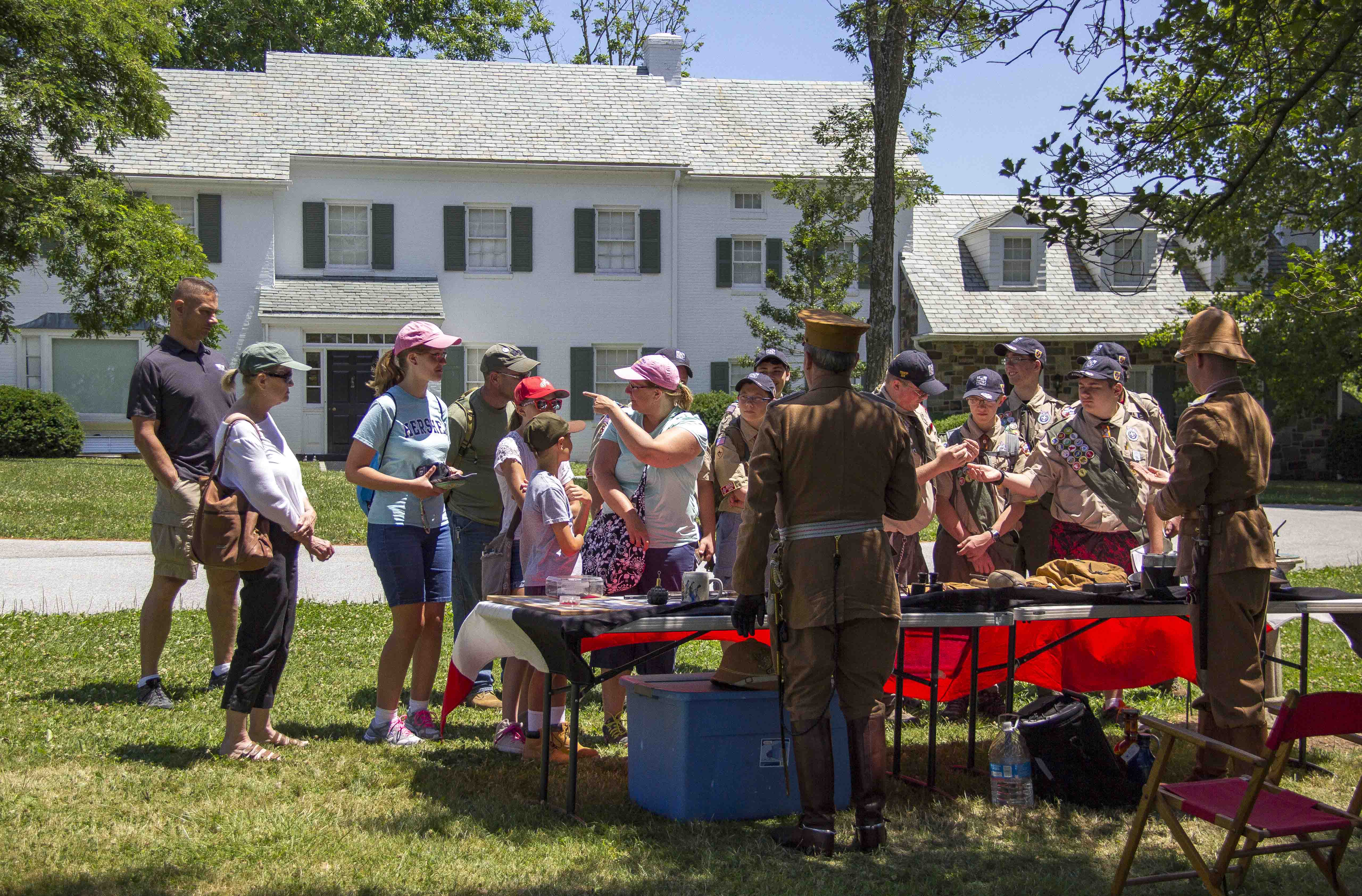 Two men dressed in khaki colored World War One uniforms stand in front of a table with a red tablecloth. On the other side of the table is a large group of boy scouts and scout leaders. Beyond is a two-story, white-sided house with green shutters.