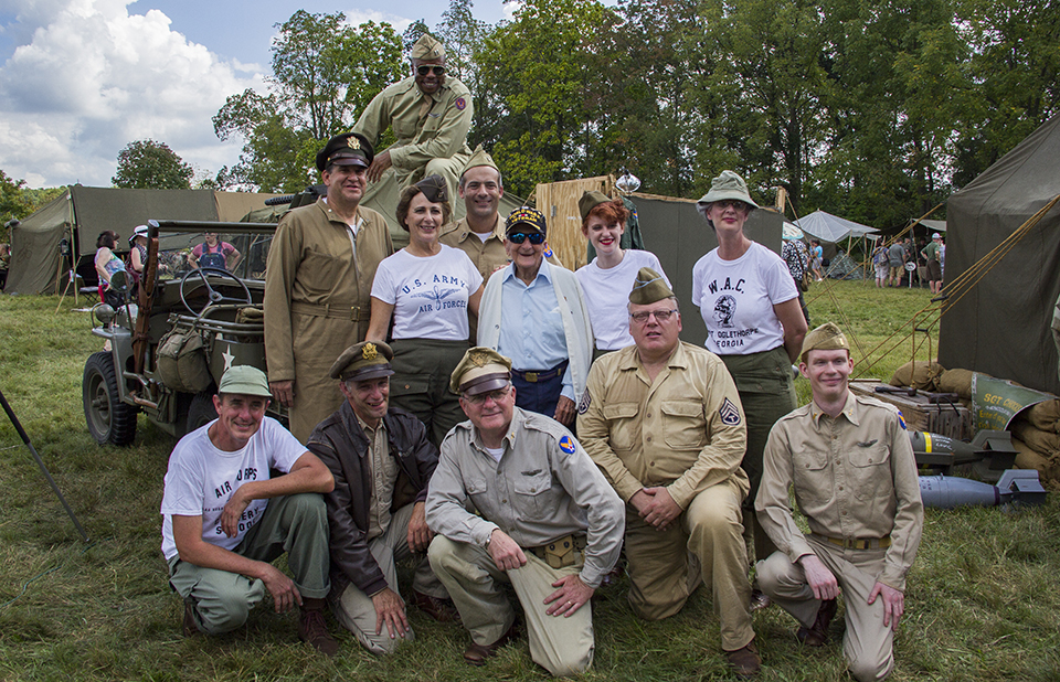 A group of World War II living historians pose for a picture with a World War II veteran. They are gathered around Jeep and camp tents are all around the group. A group of World War II living historians pose for a picture with a World War II veteran. They are gathered around Jeep and camp tents are all around the group.