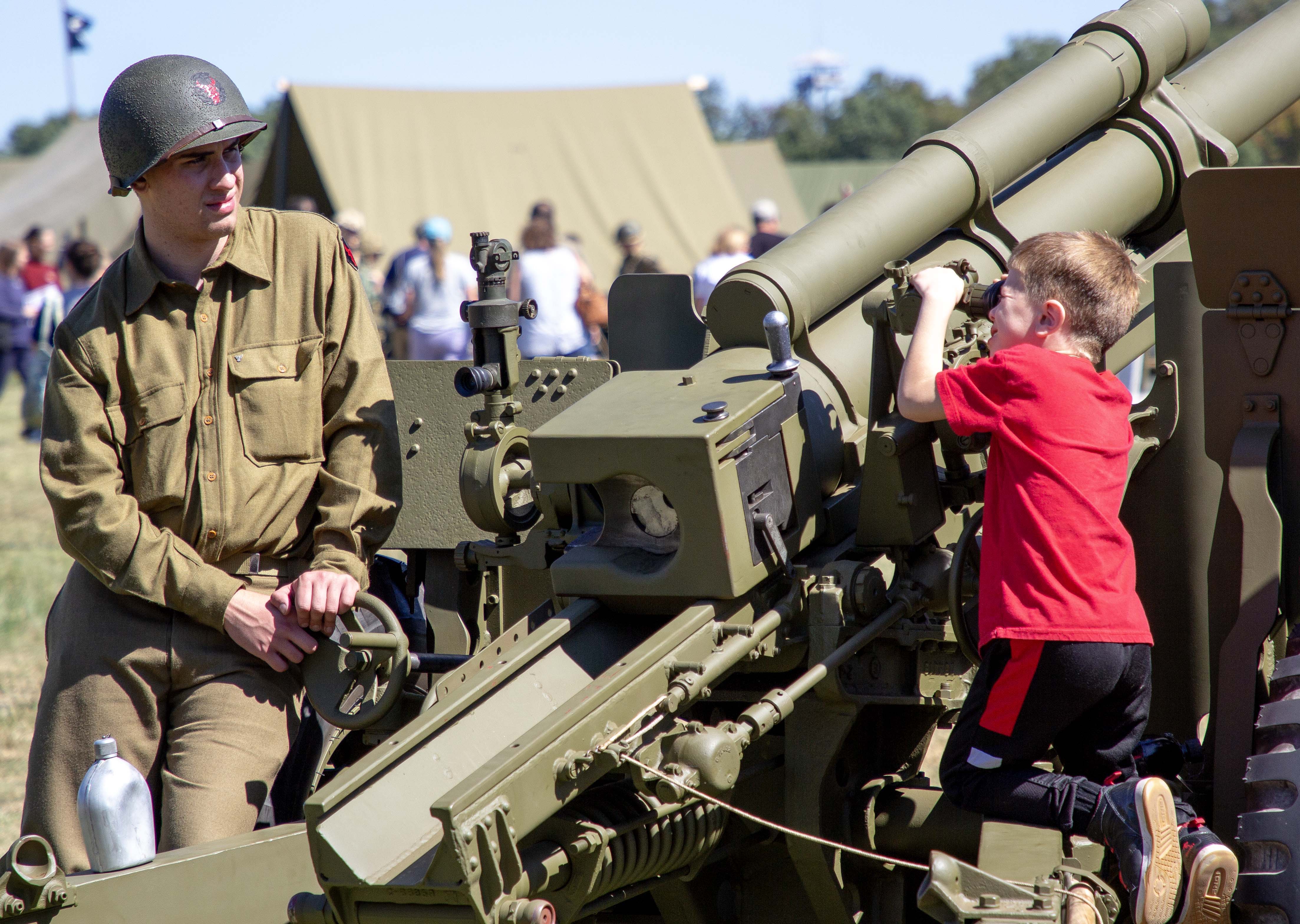 On the left a young man in full military uniform sits next to a large WWII era vehicle mounted gun while a young boy in a red shirt looks through the eyepiece.