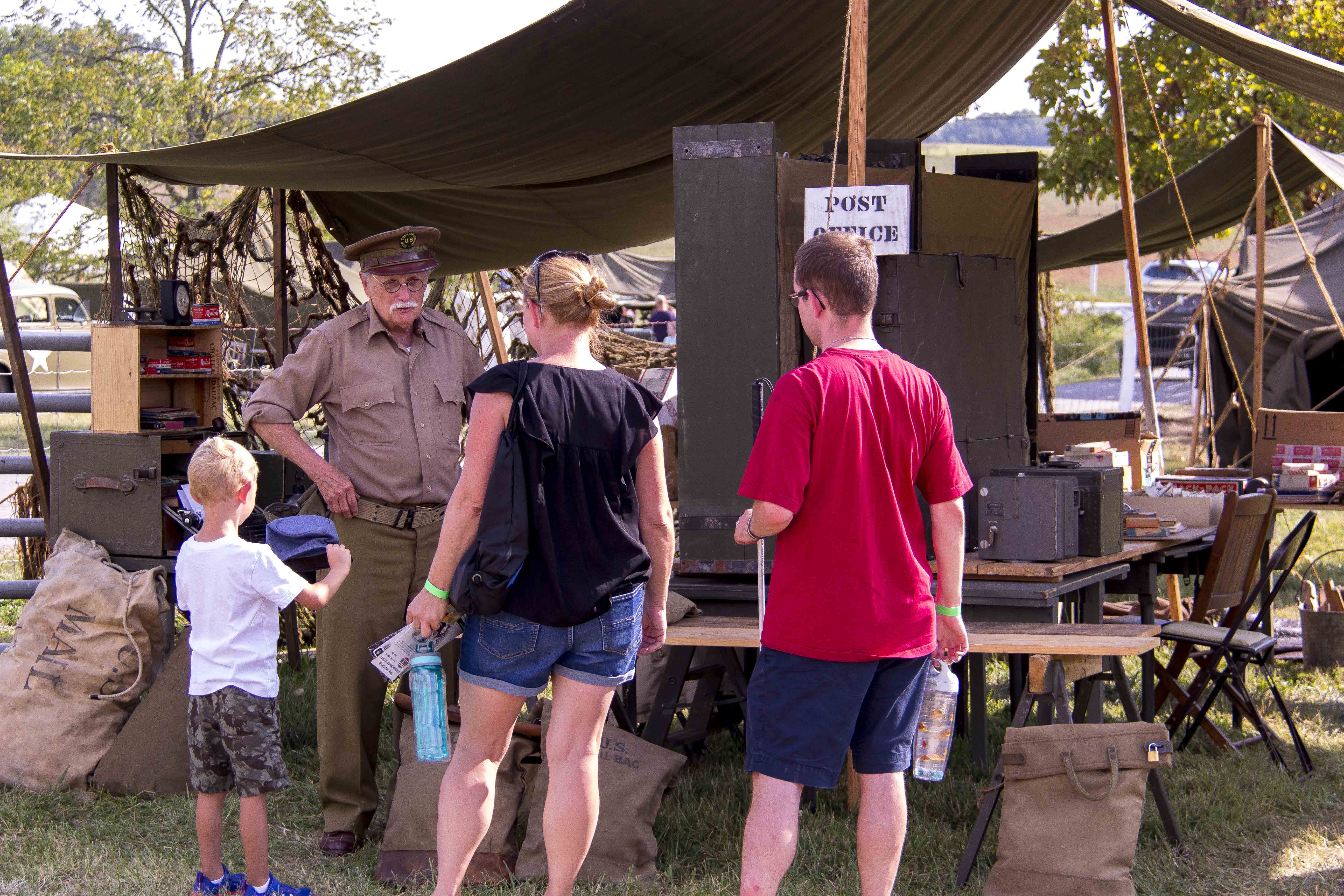 A family of three talks with a World War II living historian at his Post Office tent. There are tables with numerous boxes and shelves stack on top, full burlap mail bags sit on the ground.