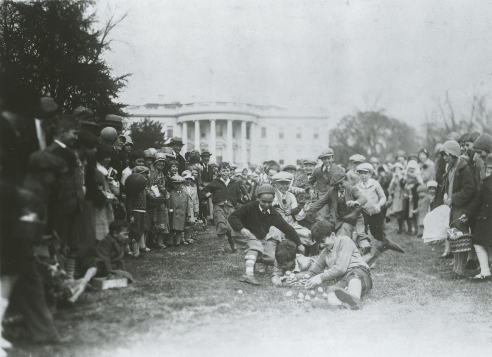An old black and white photo of a group of young children running towards the center of the photo to gather Easter Eggs. The White House is in the background and a large group of onlookers surrounds the children. An old black and white photo of a group of young children running towards the center of the photo to gather Easter Eggs. The White House is in the background and a large group of onlookers surrounds the children.