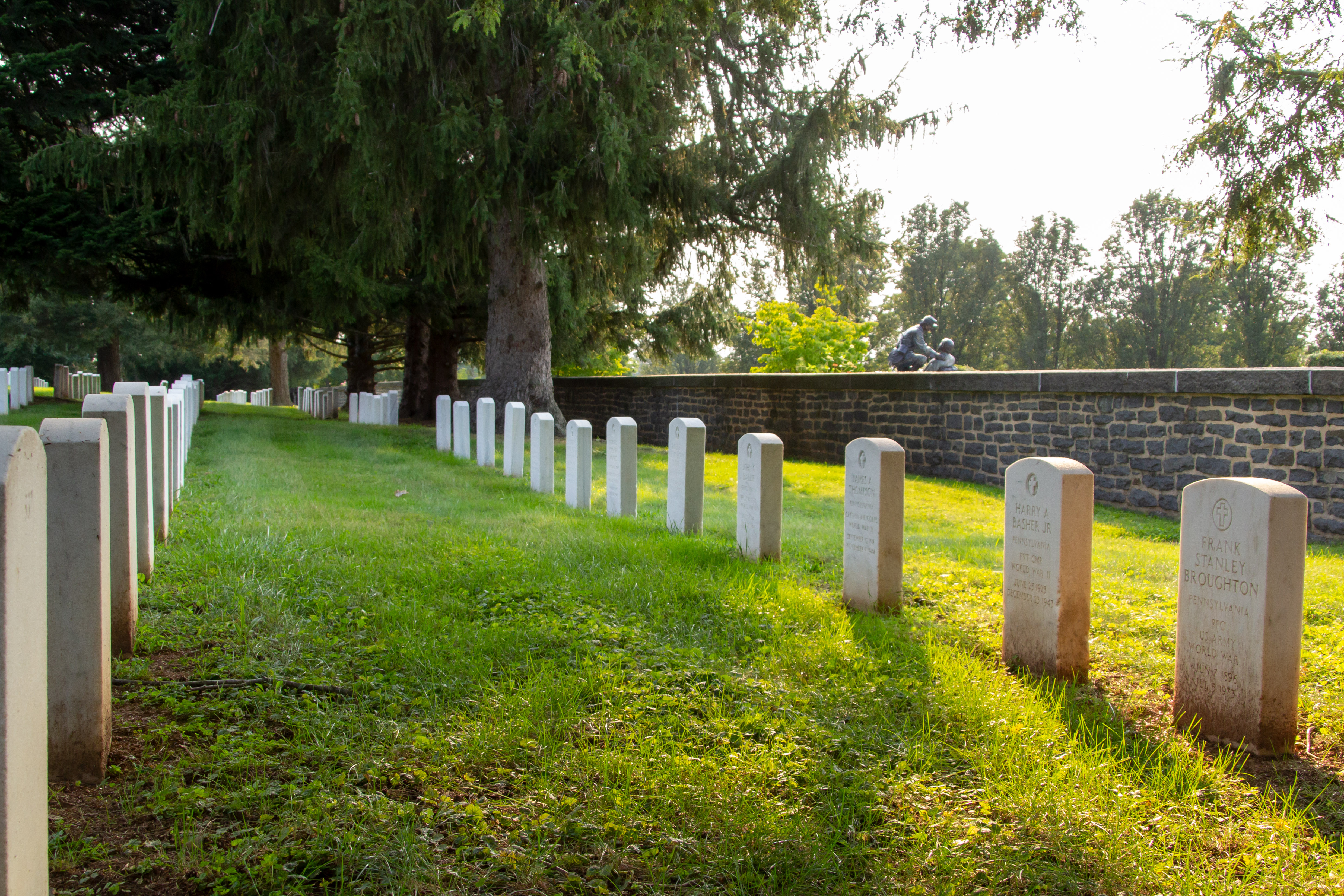 Eisenhower National Historic Site will commemorate the 80th anniversary of VE Day with special tours of Gettysburg National Cemetery. Two rows of white marble gravestones form parallel lines along a dark stone wall in a cemetery. Many mature trees are in the distance.