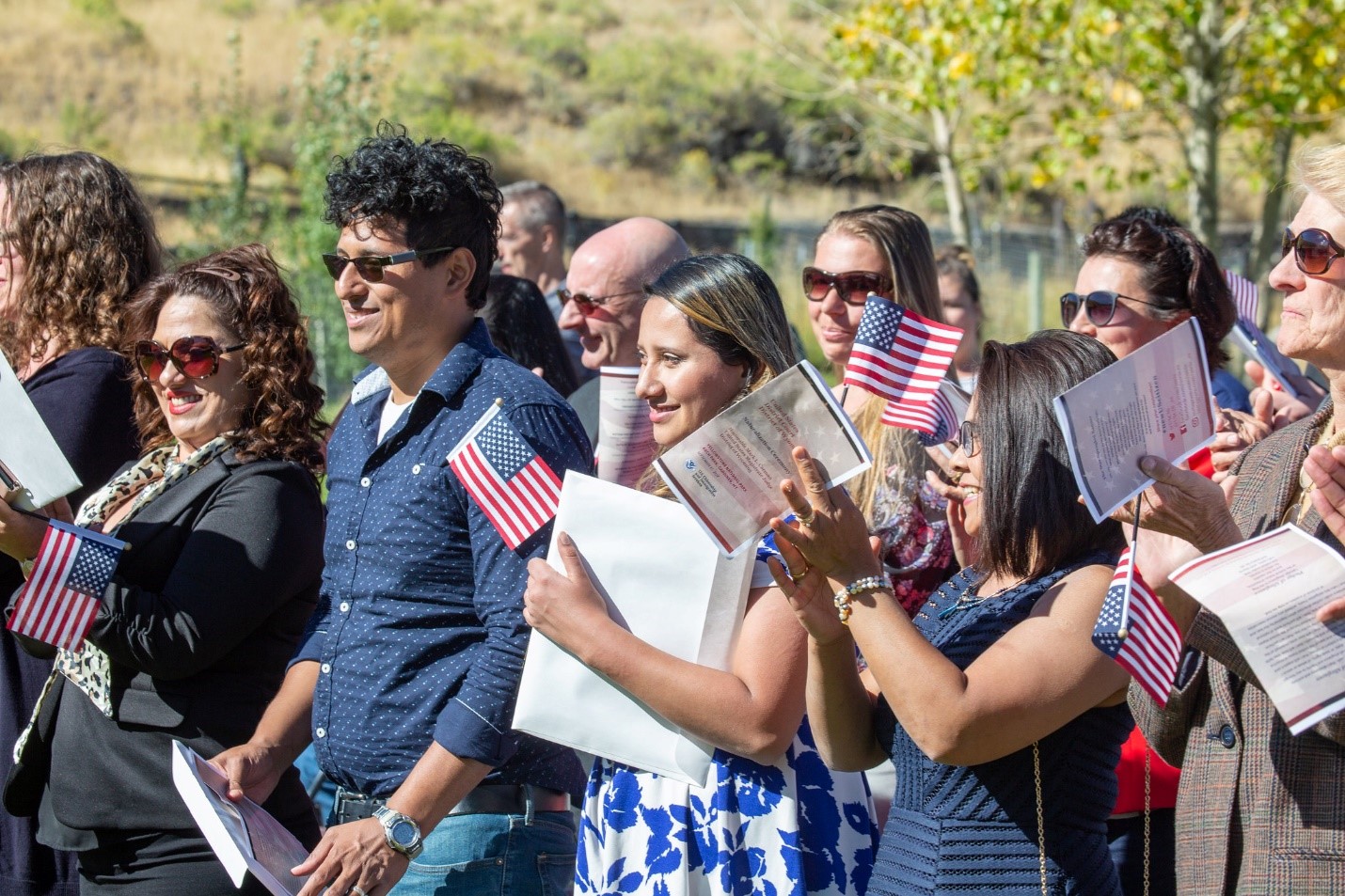 A group of people stand and hold small American flags and certificates. It's a sunny day as many are wearing sunglasses.