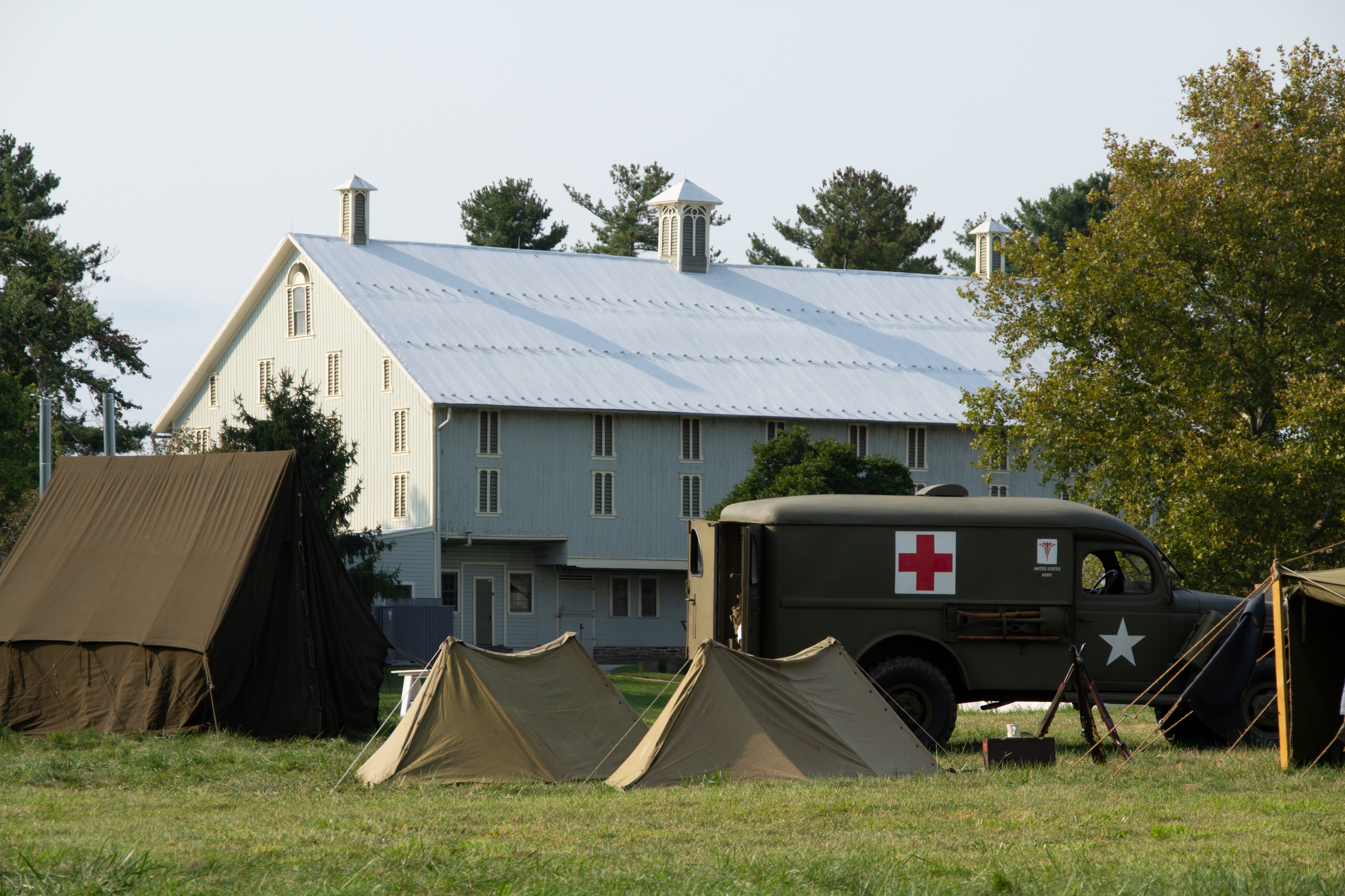 A large white barn stands in the distance with World War II tents and medical truck are in the foreground.