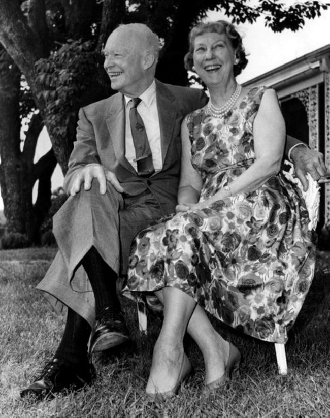 A black and white image of Dwight and Mamie Eisenhower seated and smiling behind their Gettysburg home
