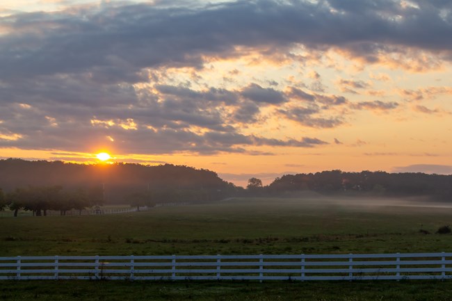 A colorful image showing the sunrise at Eisenhower National Historic Site, with rays of yellow sun spreading out over a green and brown farm field
