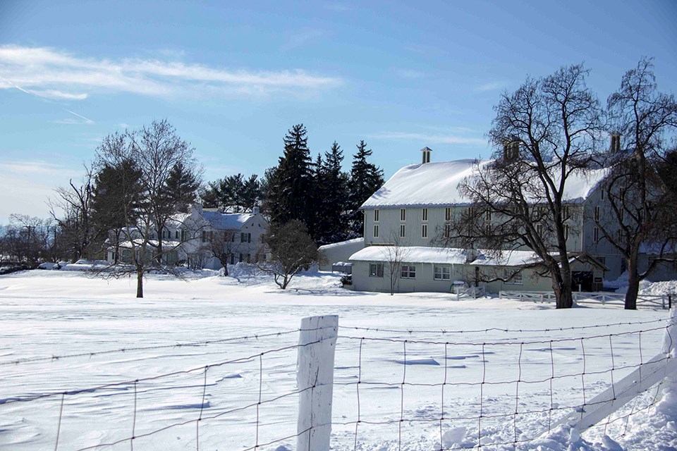 A snow covered ground with a white house and barn are nestled among trees in the distance. A wire fence is in the foreground.
