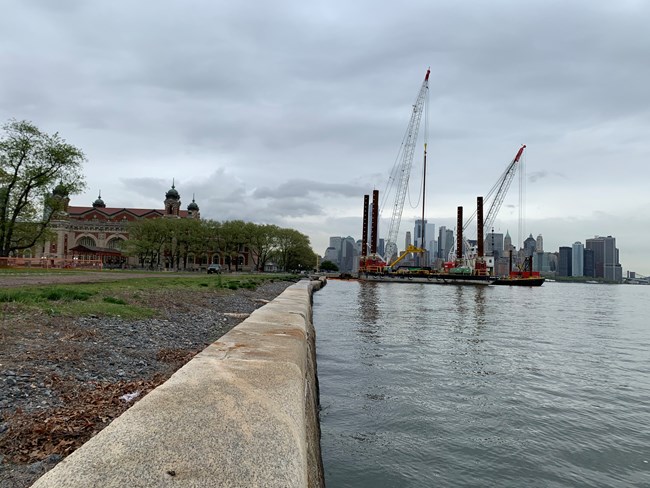 Construction on Ellis Island as seen from the south side of the island. The seawall is seen in the foreground, in the background are construction cranes and the main building of Ellis Island.