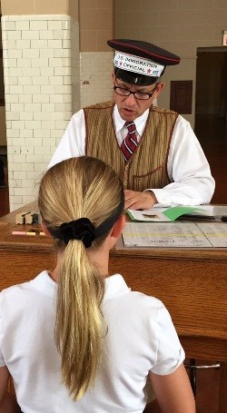 Man dressed as inspector behind desk with student role-playing an immigrant answering questions.