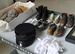 Teddy Bear, Inspecotor's hat, five pair small children's shoes and pile of white gloves sit on a table top