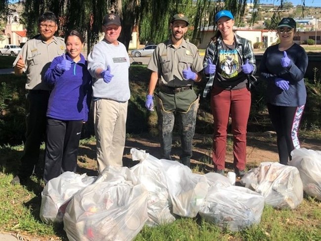 A combination of volunteers and NPS rangers stand behind a line of filled garbage bags with big smiles and thumbs up.