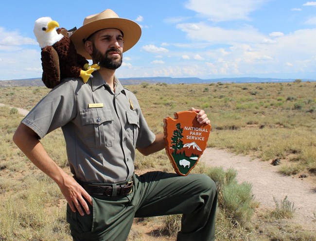 A park ranger strikes a stoic pose with a plush bald eagle and wooden National Park Service arrowhead.