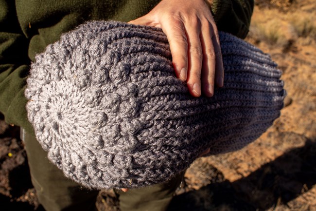 A park ranger holds a crocheted cylinder pillow in both hands.  Only the ranger's hands and torso are visible.  Focusing on the pillow, the center is relatively flat with rounded bumps.  The longer sides show crocheted ridges of yarn.
