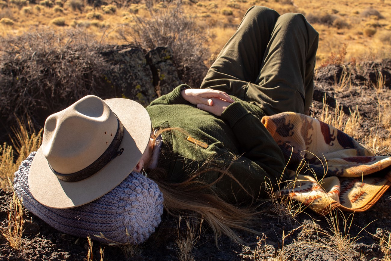 A park ranger lays down on her back on black rock with arms folded over her stomach.  Her head rests on a gray-scale crocheted cylinder pillow, and gray flat hat covers her face.  Dry, yellow grass covers the ground.