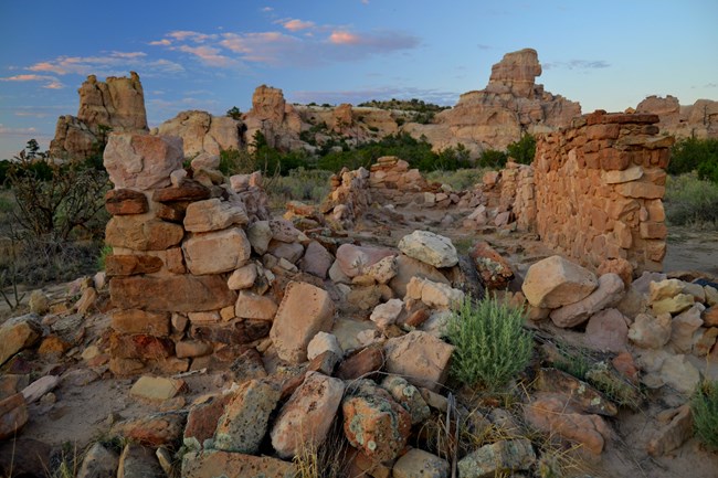 Two walls made of sandstone frame a pile of remnants of said house in between them.  Natural sandstone bluffs reach into a blue sky.