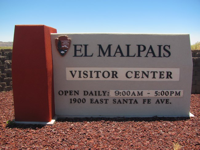 A stucco visitor center sign displays the National Park Service arrowhead, monument name, hours of operation, and street address for El Malpais National Monument.
