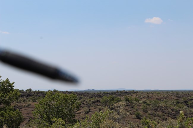 A blurred pen tip points to a distant pile of rubble that inches just above the surrounding landscape.