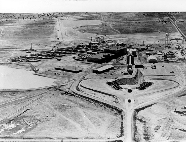 A uranium mine complex stretches across an open grassy area.  A large water basin is flanked by a small complex of single-story buildings and two larger processing structures.  (Archival photo)