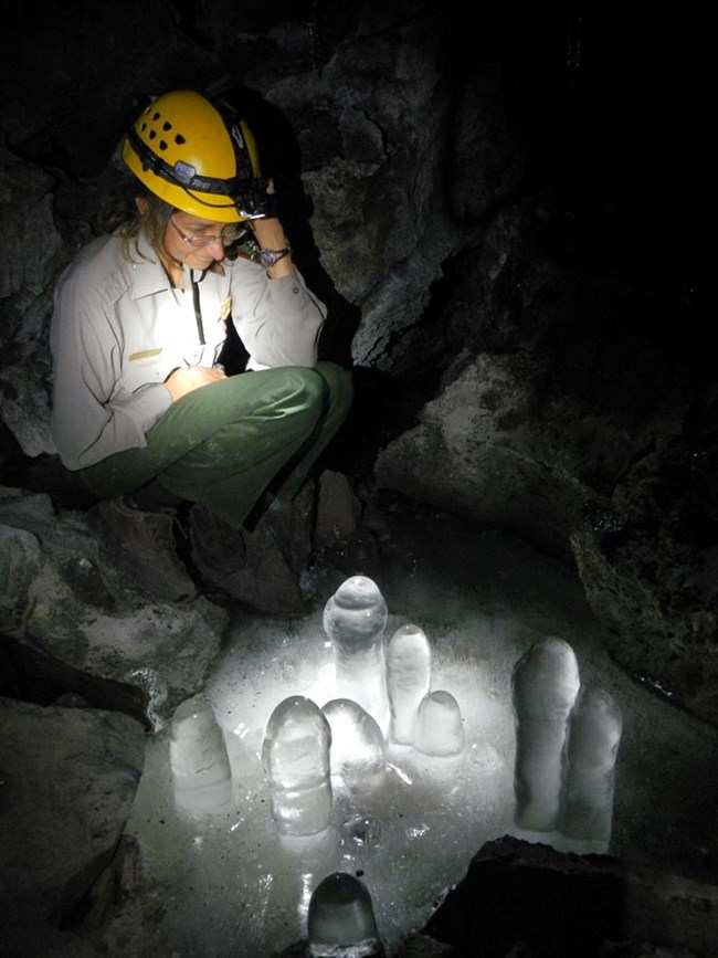 A sitting park ranger observes a frozen puddle on a cave floor.  A cluster of translucent fist-wide ice columns fill the center of the puddle.