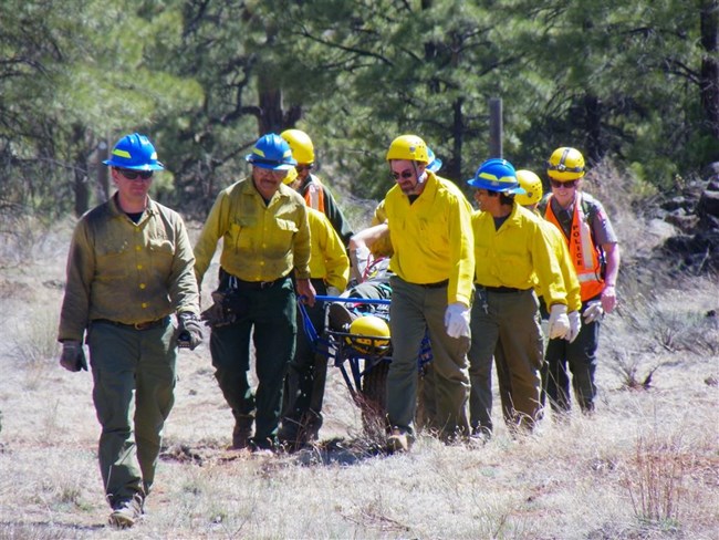 A group of people in hard hats and heavy jackets transport a practice-patient in a wheeled medical litter carry.